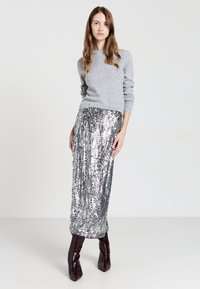 Maglione grigio a maglia con maniche lunghe abbinato a una gonna maxi di paillettes argento. Il modello indossa stivali borgogna al ginocchio. Sfondo chiaro e semplice.
