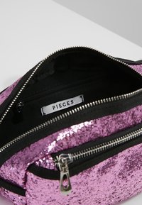 Fialová taška s flitry a černým zipem, s hlavním oddílem a vnitřní kapsou. Uvnitř je štítek s nápisem "PIECES."
