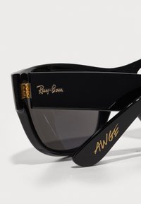 Ochelari de soare negri Ray-Ban cu un finisaj lucios, având accente aurii cu logo și lentile întunecate. Bratul este inscripționat cu 'AWGE' în litere aurii.
