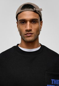 Zwarte sweatshirt met een ronde hals, voorzien van een witte kraag van een onderhemd en een blauwe grafische afbeelding op de voorzak. De pet is beige.