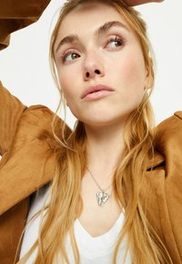 Collar plateado en forma de coraz�ón con diseño de ala de ángel, usado sobre una camiseta blanca de cuello en V, complementado por una chaqueta de ante color canela. Cabello largo, rubio y ondulado.