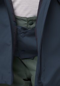 Nahaufnahme einer Person, die grüne Hosen und ein helles Hemd trägt, teilweise bedeckt von einer geöffneten dunkelblauen Jacke mit Druckknöpfen.