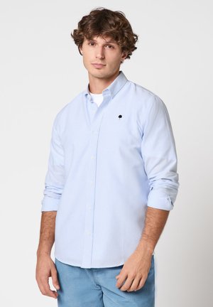 Jonge man met krullend haar, gekleed in een lichtblauw gestreept overhemd en blauwe broek, tegen een effen witte achtergrond.