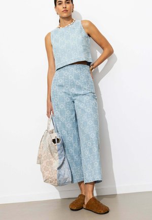 Femme portant un crop top bleu clair sans manches à motifs et un pantalon large assorti, tenant un grand sac fourre-tout patchwork et des chaussures marron sans lacets.
