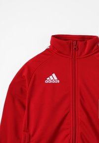 Röd Adidas sportjacka med vit logotyp på bröstet, blixtlås fram och hög krage, utställd platt på en vit bakgrund.