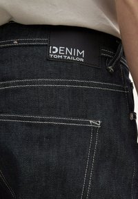 Jeans de mezclilla oscura con una etiqueta negra que lleva el texto blanco "DENIM TOM TAILOR". Costuras visibles y bolsillos en la parte trasera.