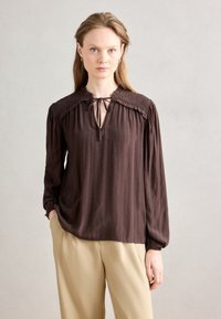 Brun langærmet bluse med nøglehulshalsudsk�æring, smockede skuldre og struktureret stof. Kombineret med beige bukser.
