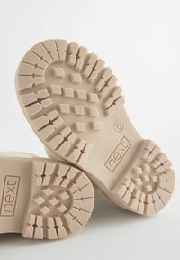 Semelles de bottes en caoutchouc beige clair avec une semelle extérieure texturée et à motifs. Comprend des rainures profondes et de petits détails carrés. Logo de la marque visible.