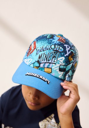 Jongen draagt een blauwe Minecraft-pet met de tekst "Diamond Miner!" en gepixeleerde grafische ontwerpen, past de pet aan met zijn rechterhand.