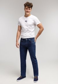 Biała bawełniana koszulka z małym niebieskim logo, w połączeniu z ciemnoniebieskimi dżinsami. Model ma na sobie granatowe tenisówki z brązowymi sznurówkami.