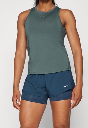 Sportliche Person trägt ärmelloses, dunkelgrünes Sporttop und marineblaue, geschichtete Shorts mit Nike-Logos auf beiden Kleidungsstücken.