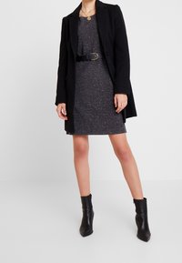 Manteau noir porté sur une robe tricotée foncée à pois, cintrée avec une large ceinture noire, associée à des bottines noires à bout pointu.