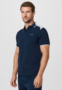 Muž v tmavomodrej a bielej krátkosleevej polokošeli s logom "Hackett", stojaci so uvoľnenými rukami proti jednoduchému svetlému pozadiu.