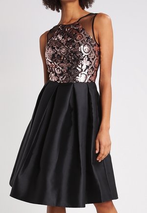 Kobieta w czarnej sukience bez rękawów z błyszczącym kwiatowym wzorem na gorsecie i rozkloszowaną plisowaną spódnicą stoi na jednolitym tle.