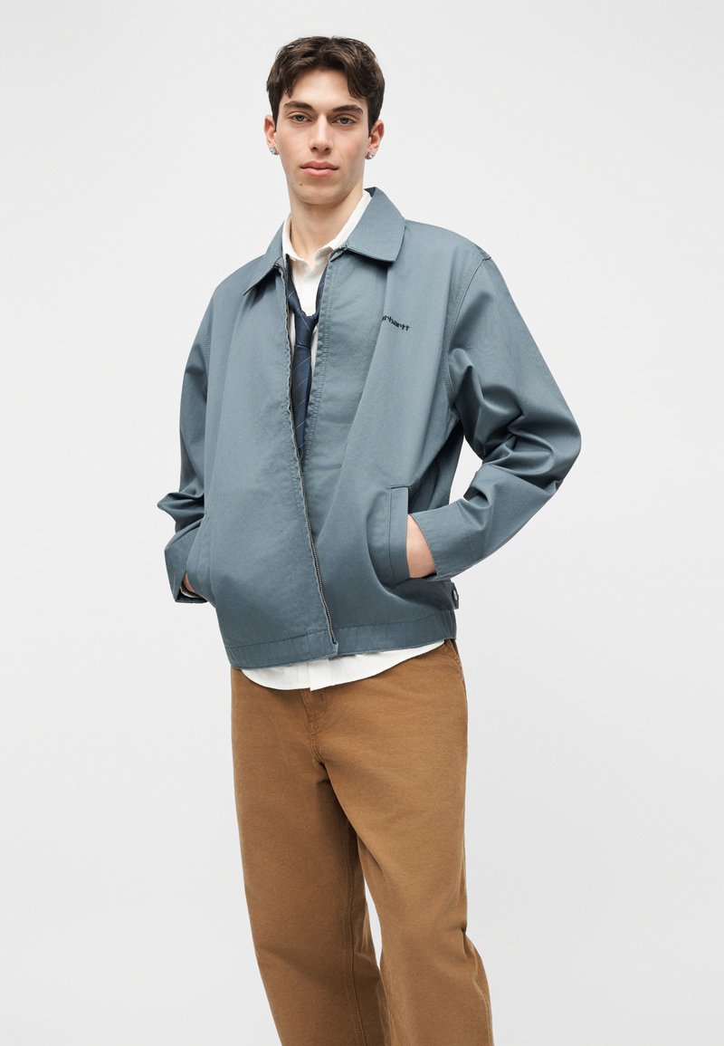 Jeune homme portant une veste bleue à fermeture éclair, une chemise blanche, une cravate bleu marine et un pantalon marron, les mains dans les poches, sur un fond uni.