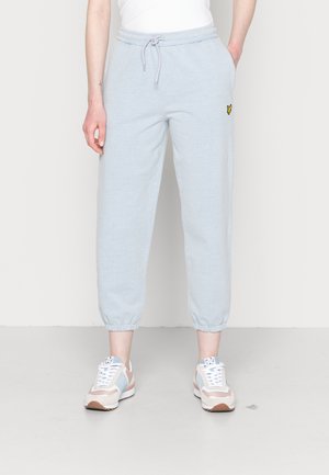 Osoba nosząca jasnoniebieskie spodnie dresowe z elastycznymi ściągaczami i małym żółtym logo na udzie, połączone z pastelowymi sneakersami.
