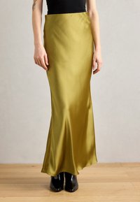 Olive green satin maxi skirt with a smooth texture, fitted at the waist and flaring slightly towards the hem. Worn with black heel boots.