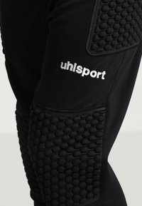 Zwarte sportbroek met textuur in de vorm van hexagonale padding op de knieën, voorzien van het 'uhlsport' logo in witte borduursel op de bovenbeen.