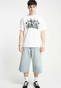 Biała koszulka z czarnym wzorem graficznym przedstawiającym psy oraz słowo "BEZLĘKOWY". Połączona z jasnoniebieskimi dżinsowymi szortami i czarnymi trampkami.