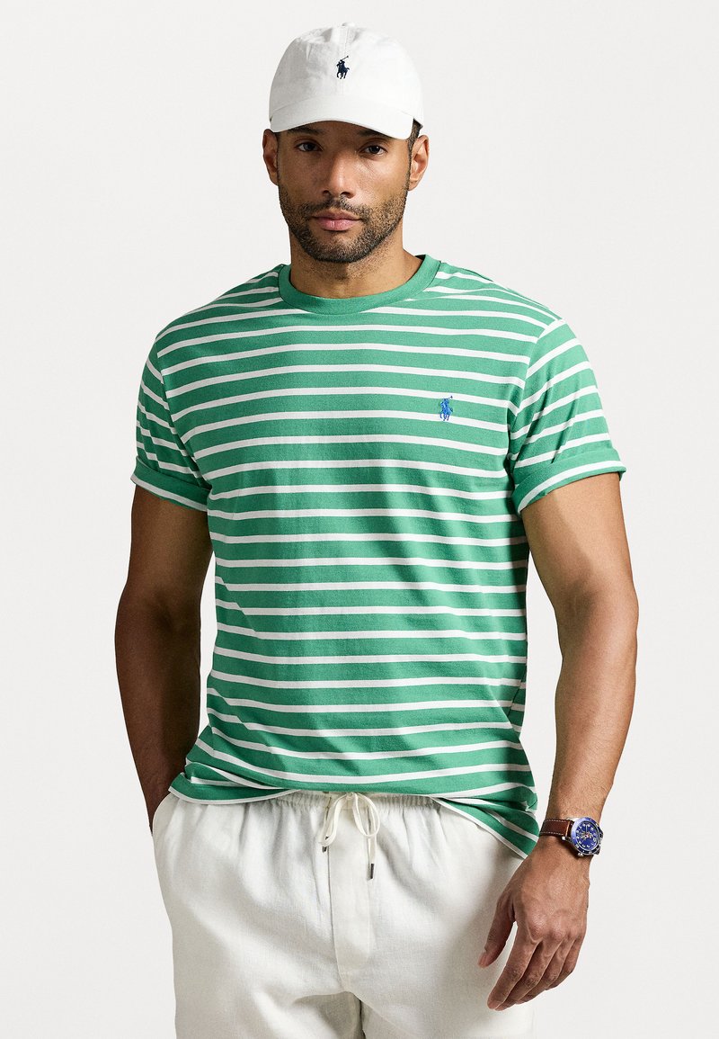 Tricou cu dungi verzi și albe, cu mâneci scurte, din material din bumbac, având un mic logo albastru. Asortat cu pantaloni scurți de culoare deschisă și o pălărie albă.