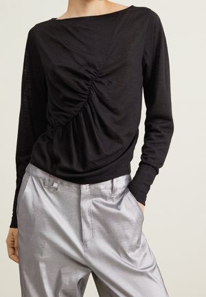 Musta pitkähihainen toppi, jossa on diagonaalinen rypytetty yksityiskohta, yhdistettynä löysiin hopeisiin satiinihousuihin, malli seisoo käsi taskussa.