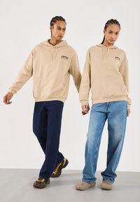 Deux personnes portant des sweat-shirts beiges et des jeans amples se tiennent côte à côte devant un fond blanc uni, toutes deux regardant droit devant.