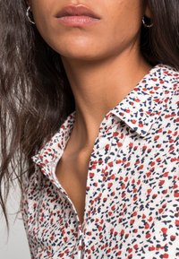 Kvindelig person iført hvid skjorte med røde og blå abstrakte pletter, sølvfarvede hoop øreringe, med langt mørkt hår synligt på venstre side.