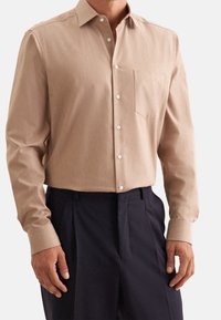 Camicia beige a maniche lunghe con colletto classico, tasca frontale e bottoni bianchi. Abbinata a pantaloni scuri, che mostrano una vestibilità su misura.