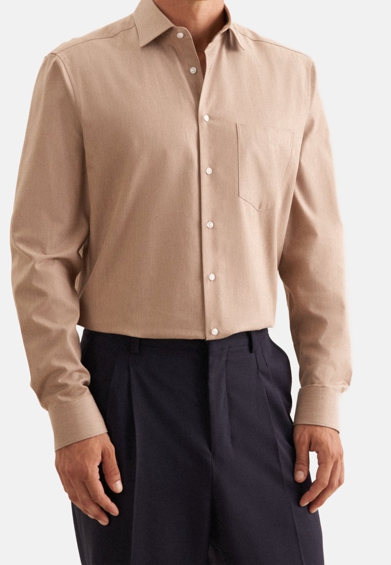 Camicia beige a maniche lunghe con colletto classico, tasca frontale e bottoni bianchi. Abbinata a pantaloni scuri, che mostrano una vestibilità su misura.