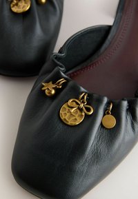 Ballerines en cuir noir avec bords froncés et breloques dorées comprenant une étoile, un disque avec un nœud, et un pendentif rond.