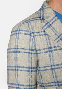 Beżowa marynarka w kratę z niebieskimi i ciemnoniebieskimi liniami. Teksturowana tkanina; widoczne detale z wyciętym kołnierzem i dopasowanym krojem.