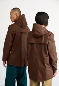 Veste de pluie marron avec capuche, dotée d'un design à évent à l'arrière. Fabriquée en matériau lisse et résistant à l'eau. Portée par deux personnes.