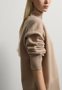 Donna che indossa un maglione beige a collo alto con le maniche arrotolate, mostrata di profilo su uno sfondo chiaro e semplice.