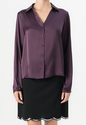 Femme portant un chemisier en satin violet à manches longues avec boutons et une jupe noire avec un bord festonné argenté.