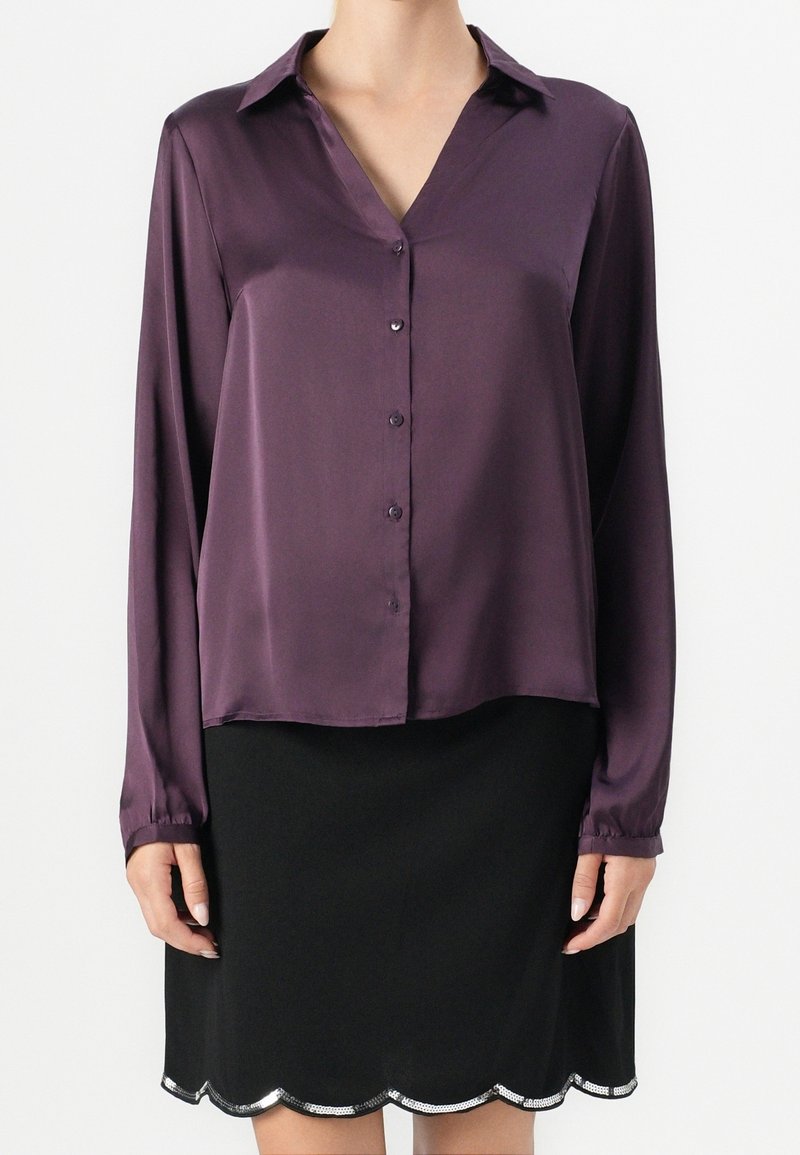 Femme portant un chemisier en satin violet à manches longues avec boutons et une jupe noire avec un bord festonné argenté.