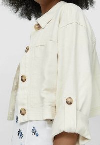 Roomkleurig spijkerjack met een cropped ontwerp, voorzien van grote bruine knopen en opgerolde mouwen. Effen textuur zonder zichtbare patronen.