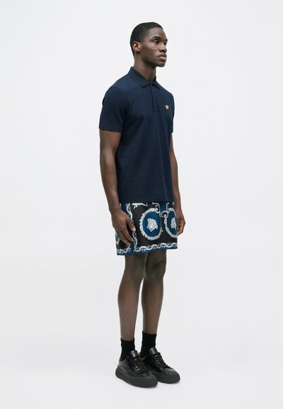 Μπλε πόλο μπλουζάκι με κοντά μανίκια και πατιλέτα με τρία κουμπιά, συνδυασμένο με εμπριμέ βερμούδα με μπλε και λευκά κυκλικά σχέδια. Μαύρα παπούτσια αθλητικού τύπου.