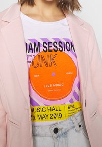 Vit t-shirt med en stor orange vinylskivgrafik, lila och gula texter, lager under en ljusrosa blazer, ihop med denimshorts.