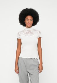 Top blanco acanalado con un forro de encaje, cuello alto y mangas cortas, caracterizado por bordes en forma de ondas y una silueta ajustada.