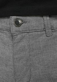 Pantalon gris texturé avec une fermeture à bouton noir et des coutures renforcées. Vue en gros plan mettant en évidence le motif du tissu et la couture de la poche.