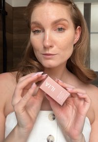 Emballage ILIA Multi-Stick, boîte matte rose pâle avec texte blanc, tenue par une personne aux cheveux longs. Fond blanc et tons de couleurs neutres.