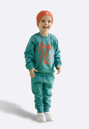 Türkiser Sweatshirt mit übergroßem orangefarbenem Text "FANTASTISCH", kombiniert mit passenden Hosen und weißen Slip-On-Schuhen, ergänzt durch eine orange Beanie.