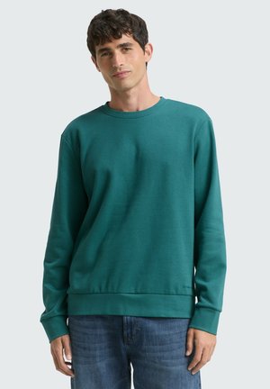 Teal sweatshirt met een ronde hals, lange mouwen en ribbels aan de mouwen. Effen textuur met een relaxte pasvorm. Gedragen over blauwe spijkerbroek.