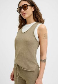 Bežový pletený top bez rukávů s kulatým výstřihem, doplněný odpovídajícími kalhotami. Outfits obsahuje texturovanou látku a detaily s tkaničkami.