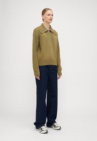 Pullover tricotado verde-oliva com colarinho com fecho, emparelhado com jeans de perna larga azul-escuro e ténis preto e branco.