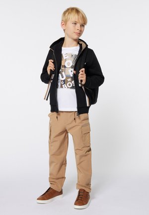 Sweat à capuche noir avec des accents rayés, T-shirt graphique blanc, pantalon cargo beige avec poches latérales et baskets brunes.