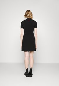 Robe noire à manches courtes, coupe ajustée, longueur au-dessus du genou, tissu lisse, avec une étiquette de logo rouge à l'arrière. Portée avec des bottines noires.