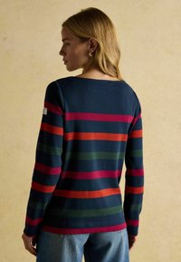 Námornícky modrý sveter s výraznými horizontálnymi pruhmi v zelenej, červenej a oranžovej farbe. Má lodičkový výstrih a priliehavé rukávy. Vyrobený z bavlnenej zmesi.