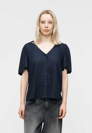 Blondynka w granatowej, krótkorękawowej koszuli na guziki i spranych czarnych dżinsach, stojąca na tle jasnego, jednolitego tła.