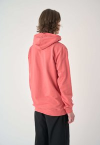 Koralfarbenes Kapuzenshirt mit Kordelzugkapuze, langen Ärmeln, gerippten Bündchen und Saum. Weiches Material, lockere Passform, Ansicht von hinten. Schwarze Hose nicht näher beschrieben.